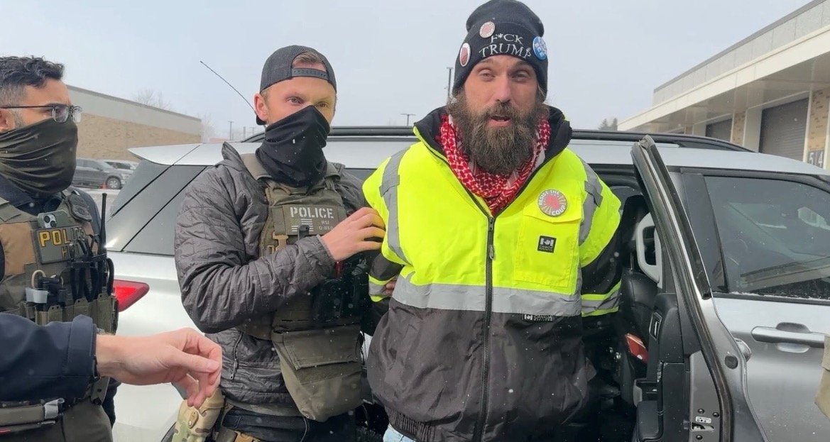 Police officers apprehend a man wearing a bright yellow jacket and a beanie with political messages during a snowy day.