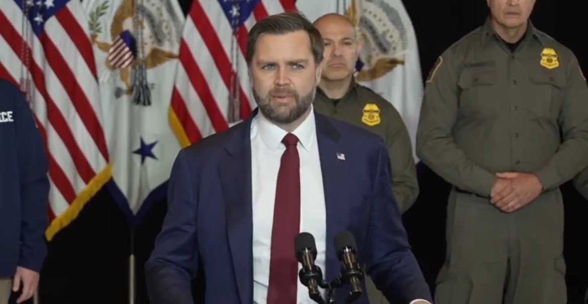 Official press conference featuring a speaker in a suit addressing an audience with American flags in the background, flanked by law enforcement personnel.