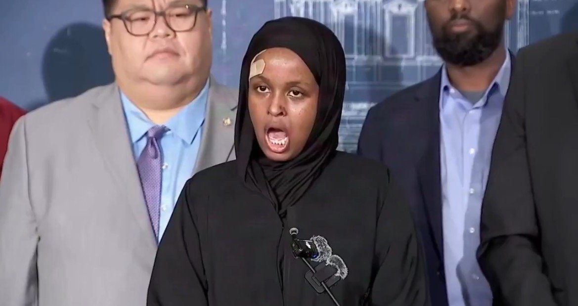 A woman in a hijab speaks at a press conference, expressing her views while flanked by three men in formal attire.