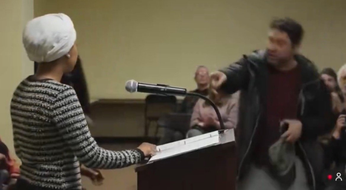 A speaker at a podium addresses an audience while a man gestures emphatically, creating a tense atmosphere during a public meeting.
