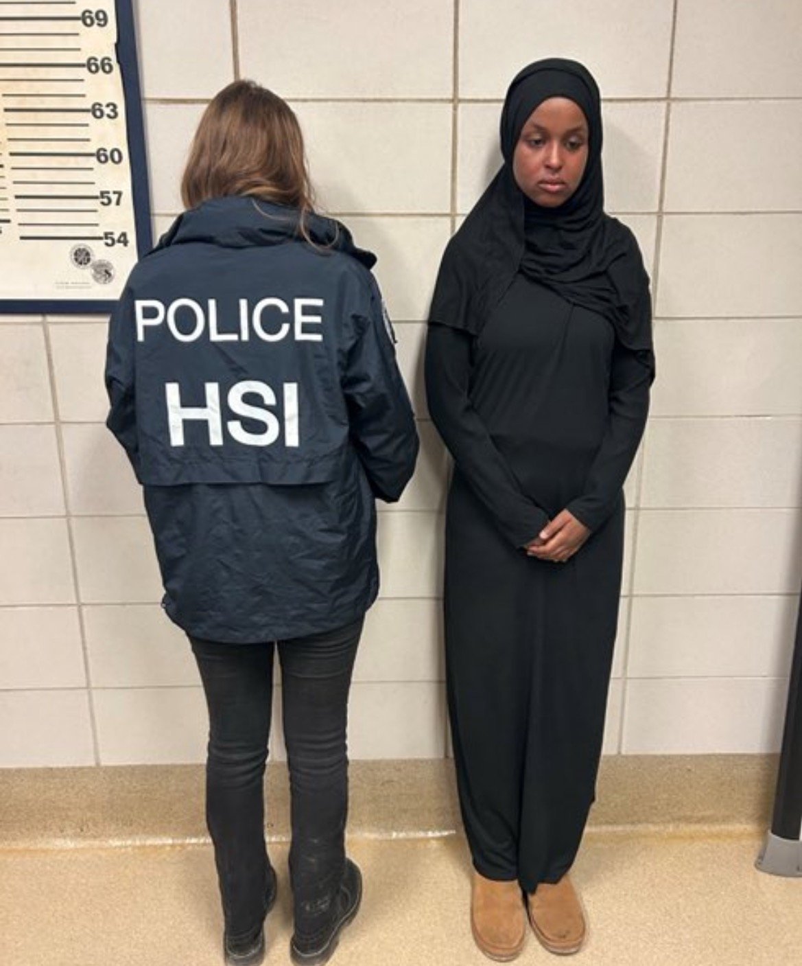 Two individuals in a police setting, one wearing a law enforcement jacket marked "HSI" and the other dressed in a black dress and hijab, standing against a wall.
