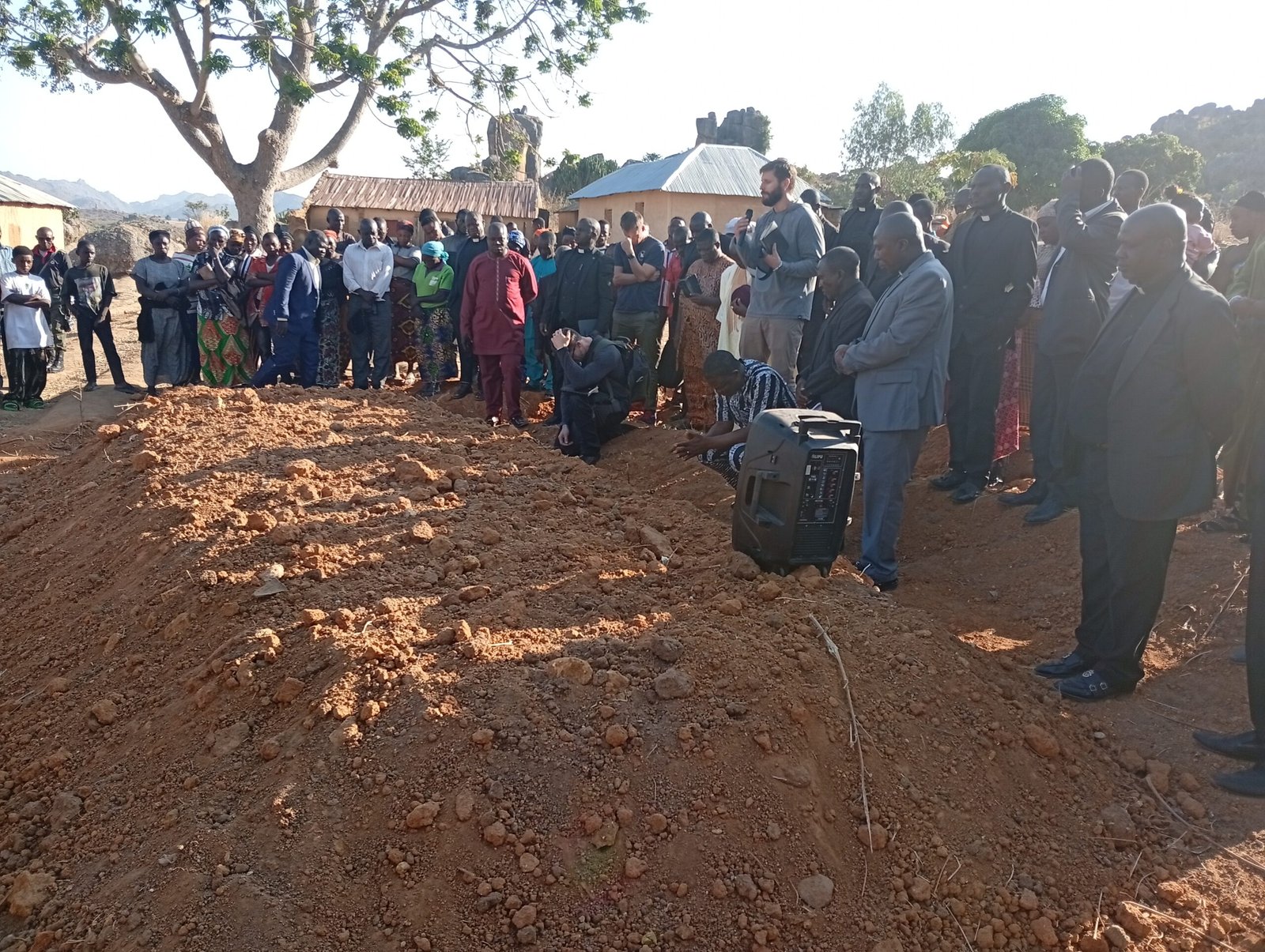 A large group of people gathered around a grave site during a solemn ceremony in a rural setting.