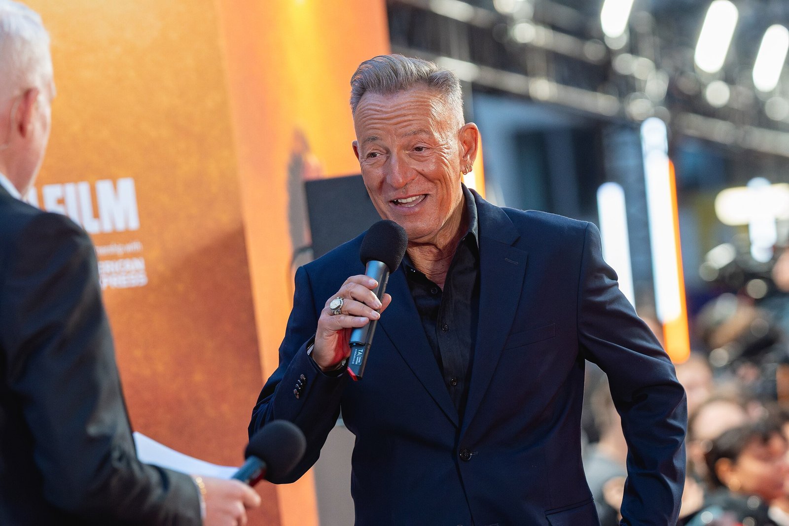 A man in a black suit speaks into a microphone at a film event, with bright lights and an audience in the background.