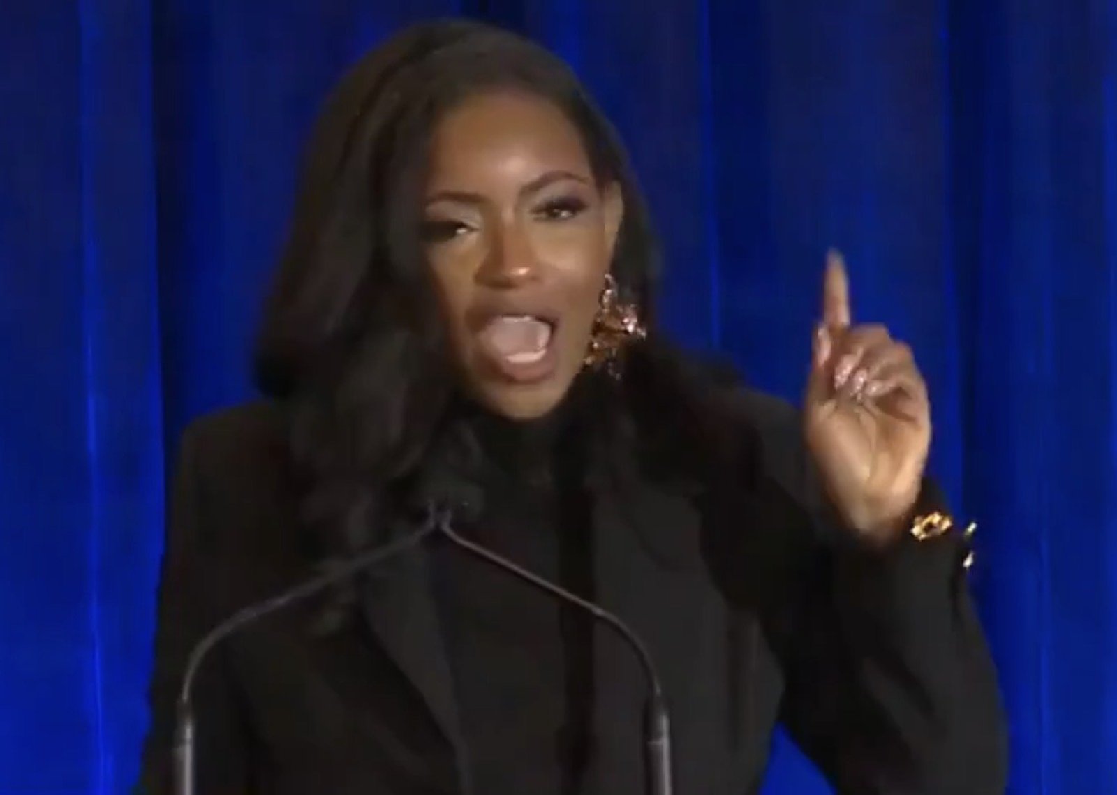 Dynamic speaker passionately addressing an audience from a podium, wearing stylish attire and dramatic earrings, with a blue backdrop enhancing the presentation.