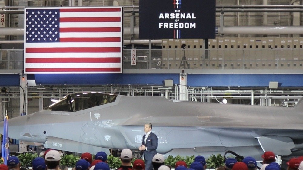 A military official speaks during the unveiling of a new fighter jet under a U.S. flag and a sign reading "The Arsenal of Freedom."