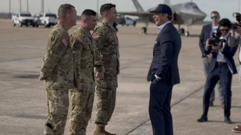 Military personnel greet a civilian in formal attire at an airfield, with aircraft and photographers in the background.