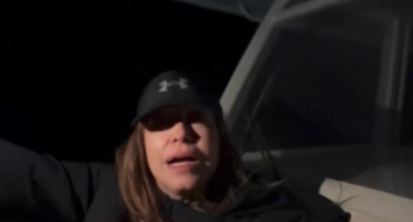 Person wearing a black hat and jacket speaks on a boat at night, with a dark background and boat structure visible.