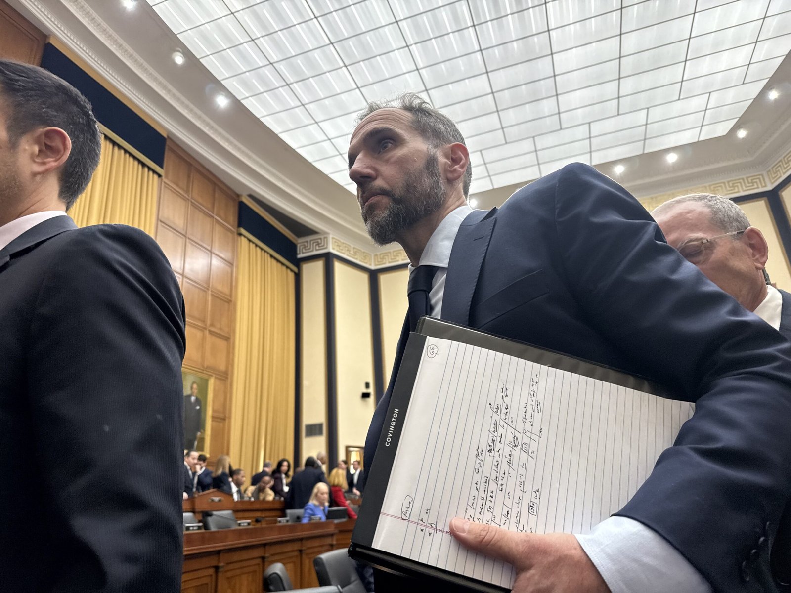 A man in a suit holds a notebook while walking through a congressional hearing room filled with people and wooden furniture.