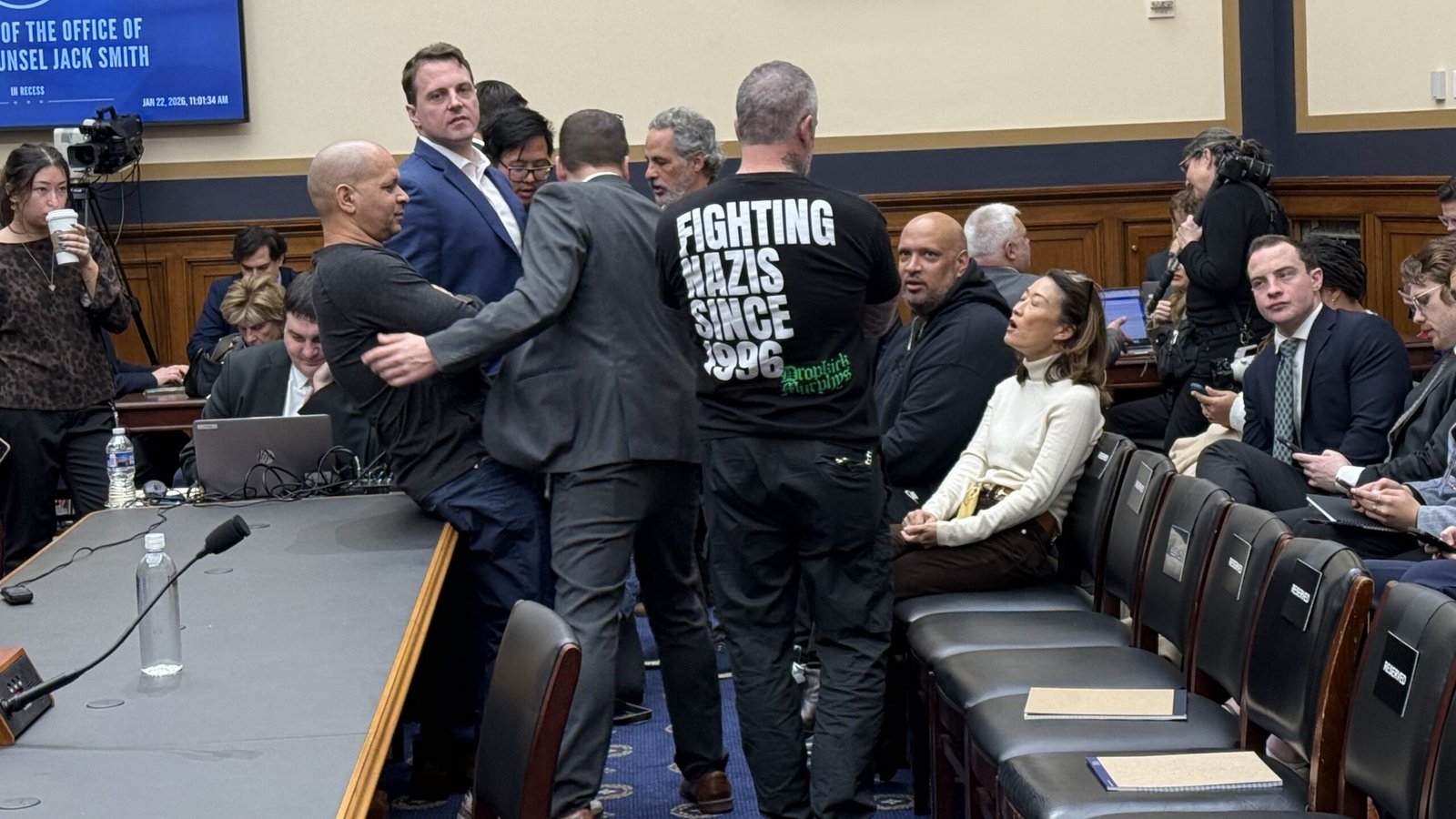 A chaotic scene in a congressional hearing room with attendees, including a man wearing a shirt that reads "Fighting Nazis Since 1996," amidst discussions and media presence.