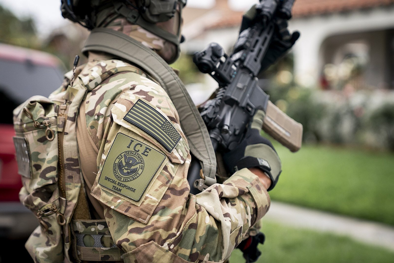 ICE Special Response Team officer in tactical gear holds a firearm, showcasing a camouflage uniform and patches, while positioned outdoors.
