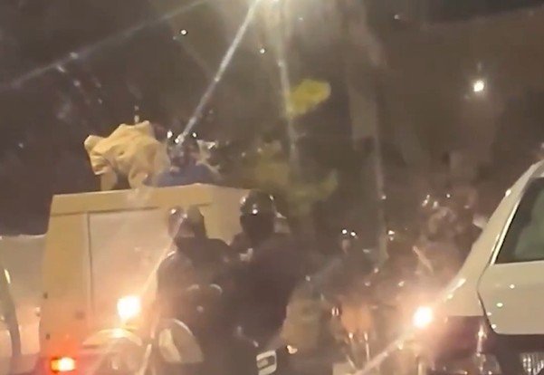 Police officers confront individuals during a nighttime disturbance, with vehicles and a crowd visible in the background.