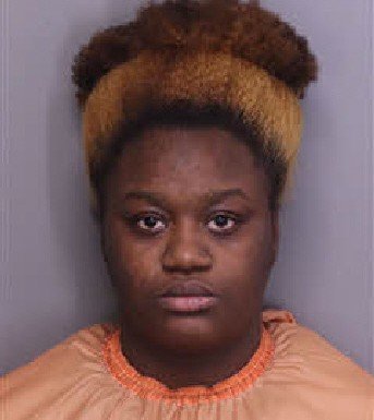 Mugshot of a young woman with curly hair wearing an orange jail uniform, looking directly at the camera.