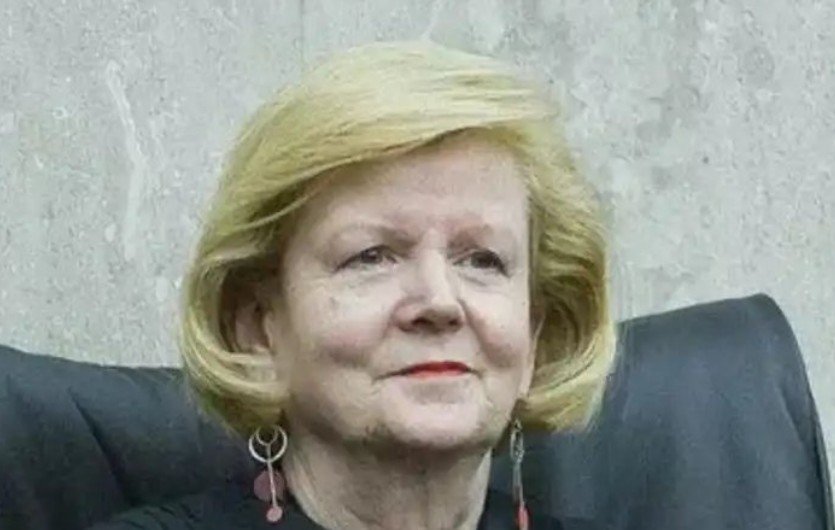 Portrait of a woman with blonde hair and earrings, seated in a formal setting, exuding a confident demeanor.