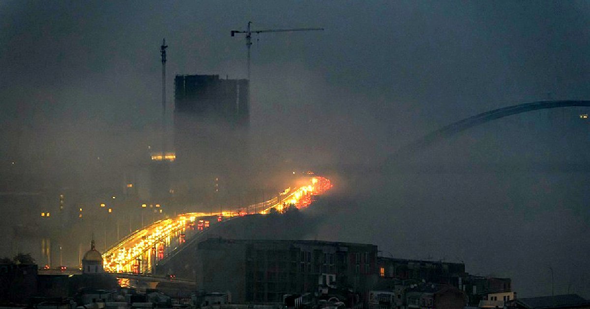 Foggy night scene of a busy bridge with headlights illuminating the road, surrounded by construction cranes and a mysterious urban skyline.