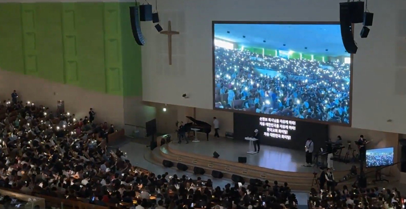 A large audience holds up lights during a worship service in a modern church setting, with a screen displaying lyrics and a speaker on stage.
