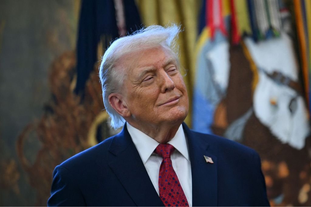 Donald Trump smiling in a formal setting, wearing a dark suit and red tie, with flags and artwork in the background.