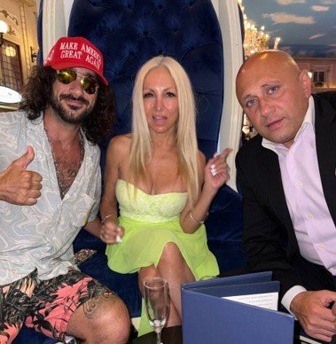 Three individuals posing together in a stylish lounge setting, with one wearing a red "Make America Great Again" cap and another in a bright yellow dress.
