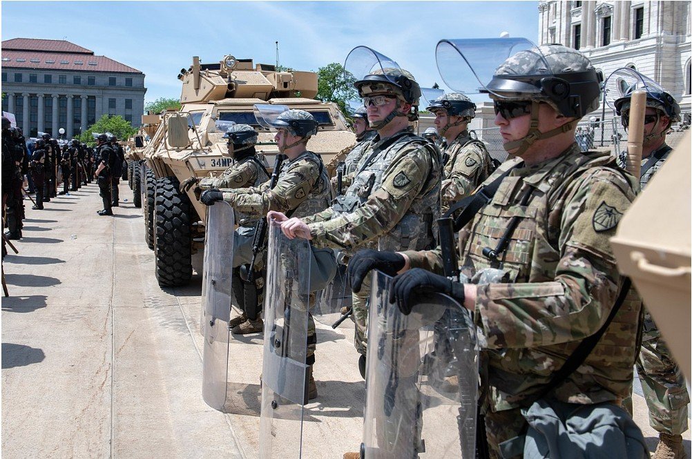 Military personnel in tactical gear stand in formation with shields and an armored vehicle during a public demonstration.