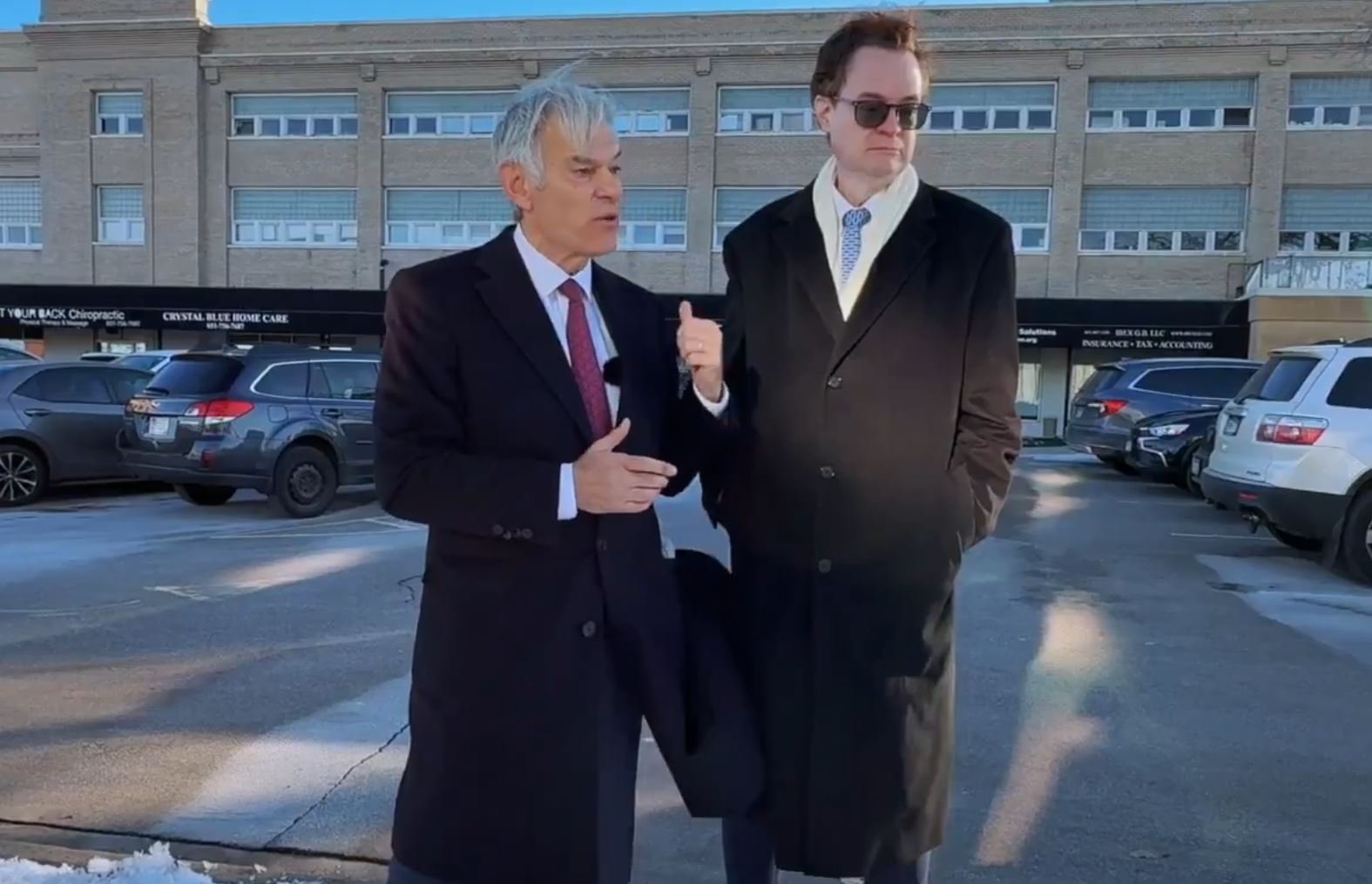 Two men in formal attire stand outdoors, discussing in front of a parking lot with various vehicles and a building in the background.