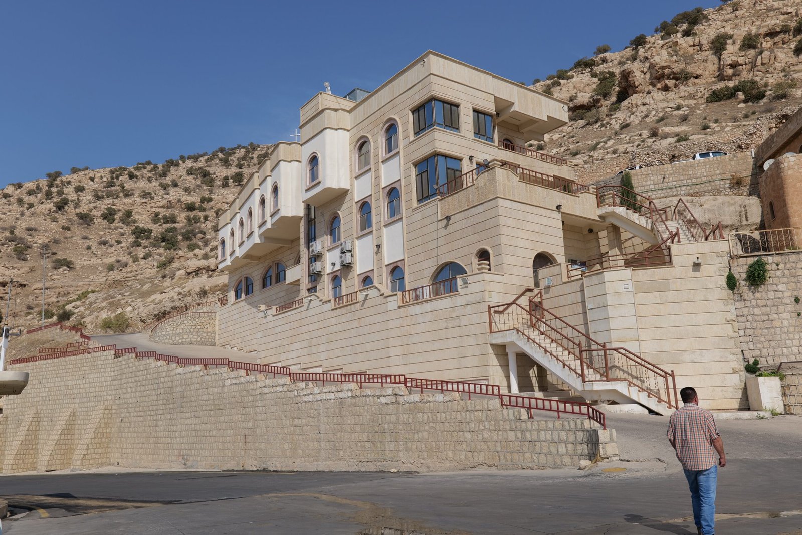 Modern building with multiple levels and large windows set against a rocky hillside, featuring staircases and a person walking nearby.