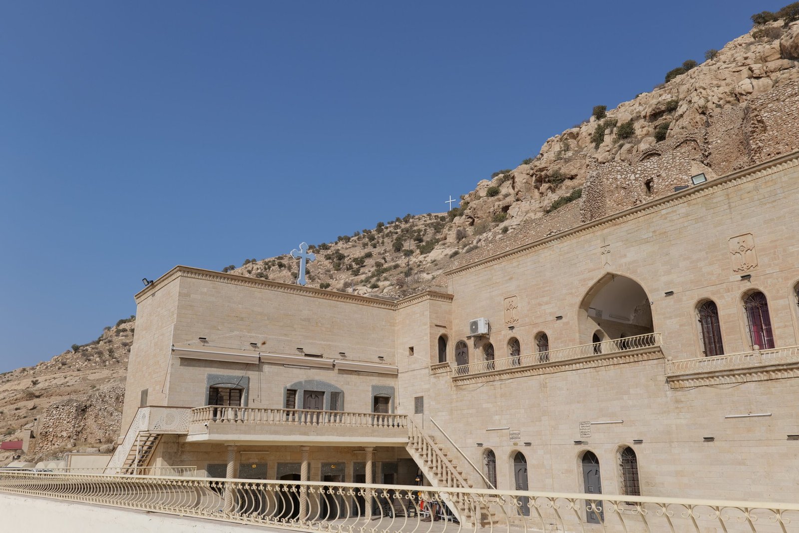 Historic stone building with arches and balconies, situated against a rocky hillside with visible crosses and a clear blue sky.