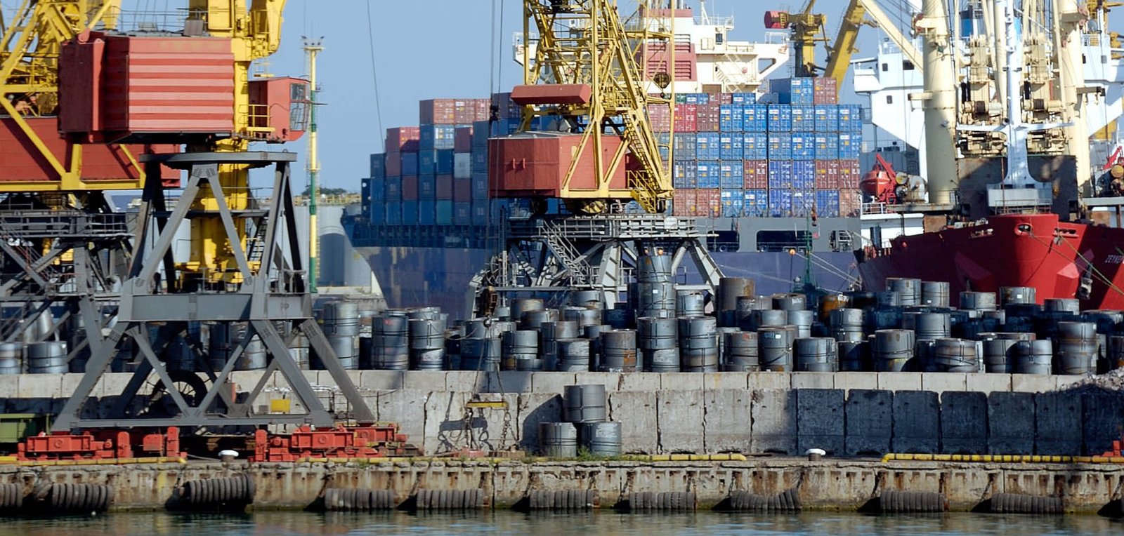 Cargo containers and barrels stacked at a busy shipping port with cranes and ships in the background, indicating maritime trade activities.