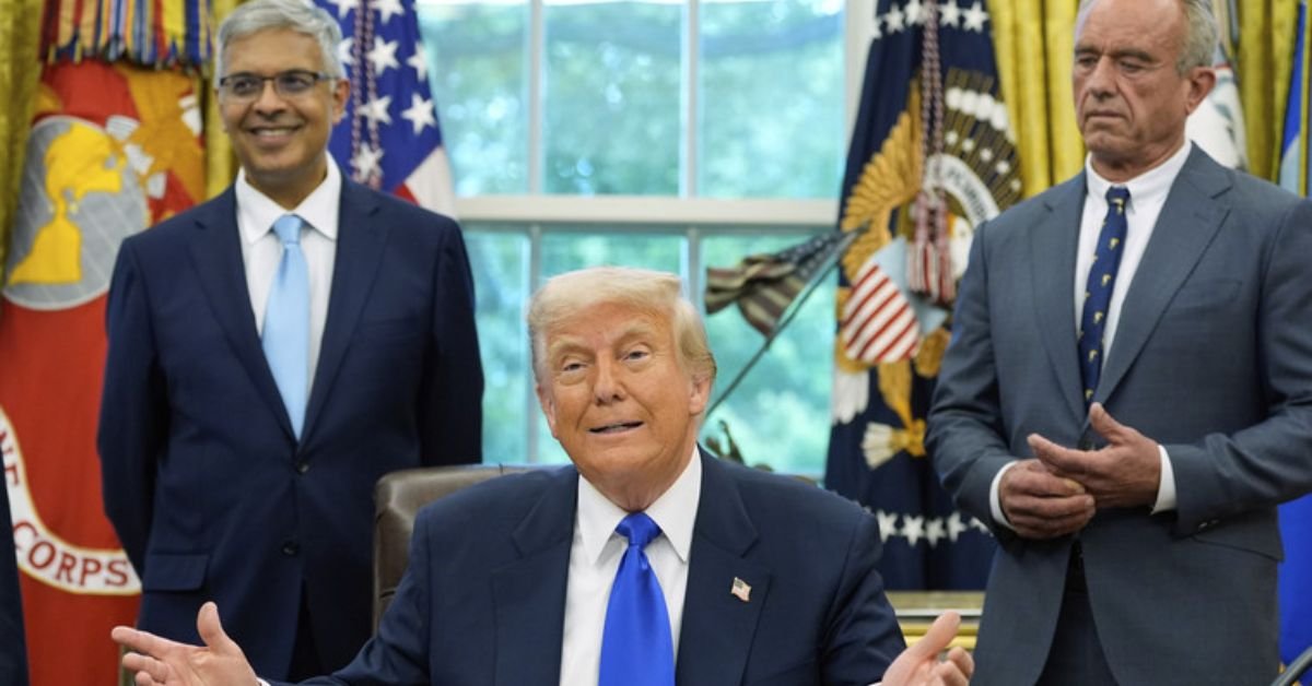 Donald Trump gestures during a meeting in the Oval Office, flanked by two men in formal attire, with presidential seals and flags in the background.