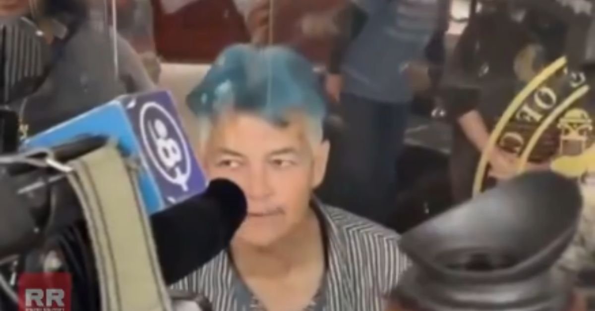 Person with blue hair sitting behind a glass barrier, surrounded by cameras and onlookers, during a media event or interview.