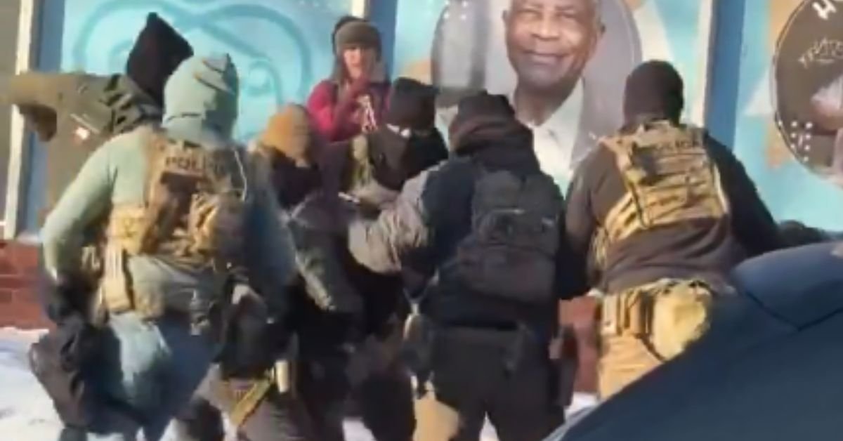 Law enforcement officers in tactical gear engage with an individual on a snowy street, with a large mural visible in the background.