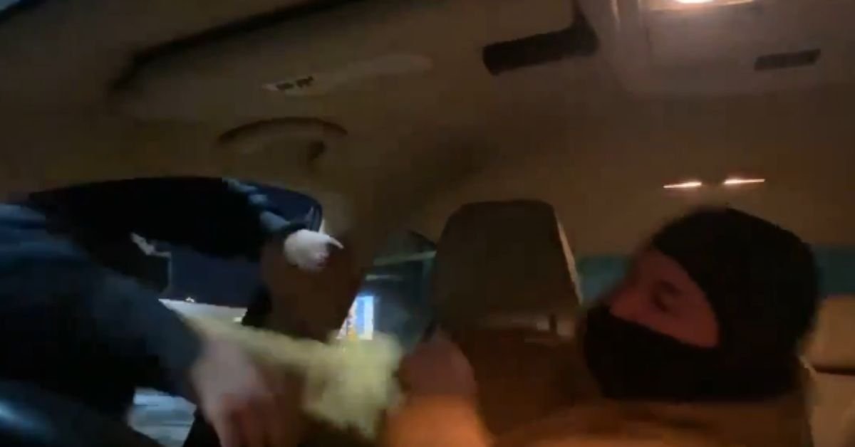 A person wearing a mask reaches into a car, while another individual seated appears surprised, capturing a tense moment inside the vehicle.