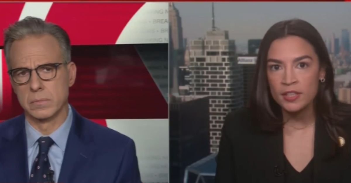 News interview featuring two political figures discussing current events against a city skyline backdrop.