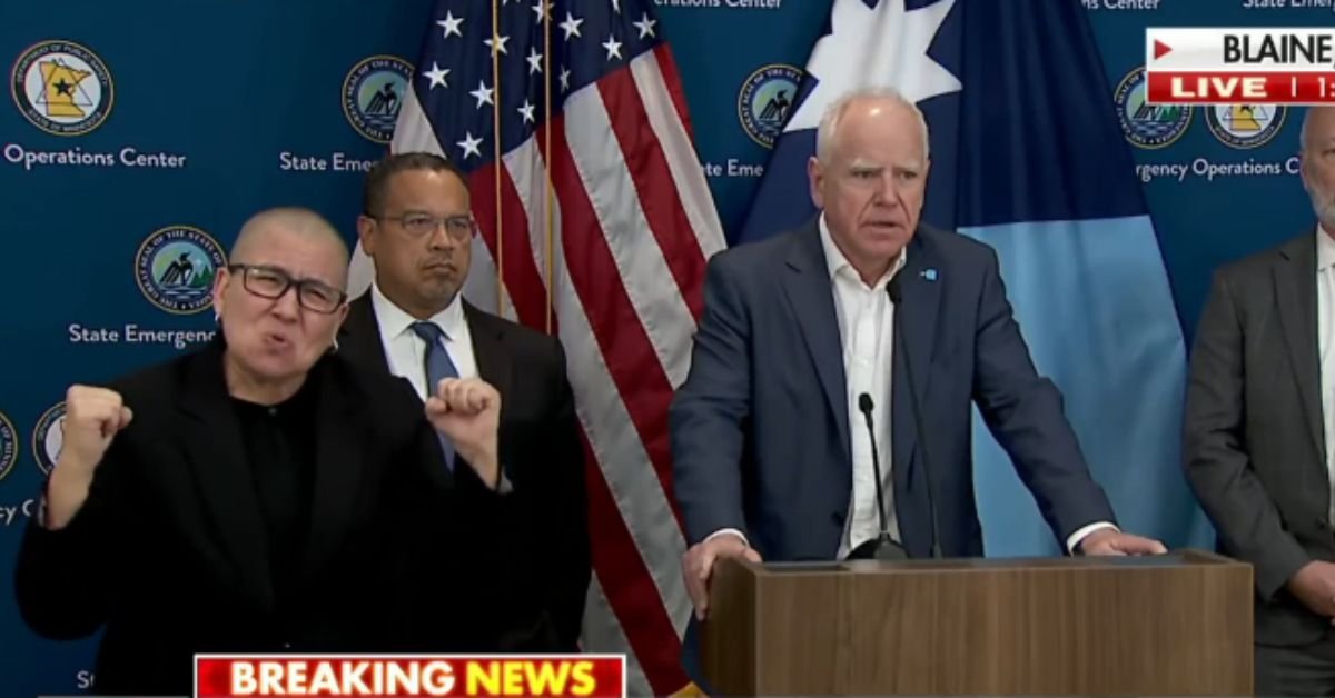 Press conference featuring state officials discussing emergency operations, with an interpreter providing sign language support, set against a backdrop of flags and emergency center logos.