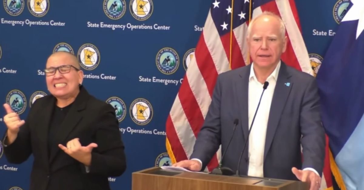 Governor speaking at a press conference with an interpreter using sign language, featuring state flags and emergency operations center branding in the background.