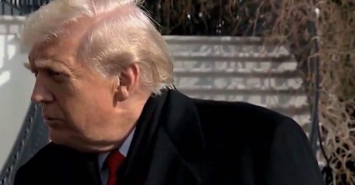 Profile view of a man with light hair and a dark coat, standing outdoors near a building, engaged in conversation.