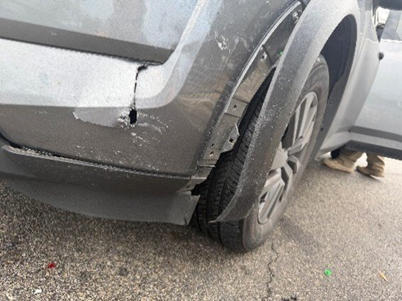 Close-up of a damaged gray vehicle showing a cracked fender and worn tire on a paved surface.