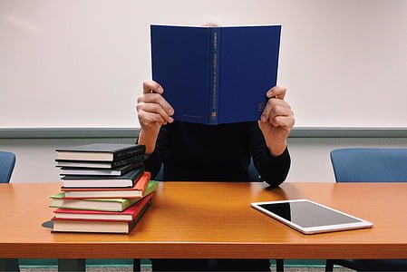 Person reading a blue book at a table with a stack of colorful books and a tablet nearby.