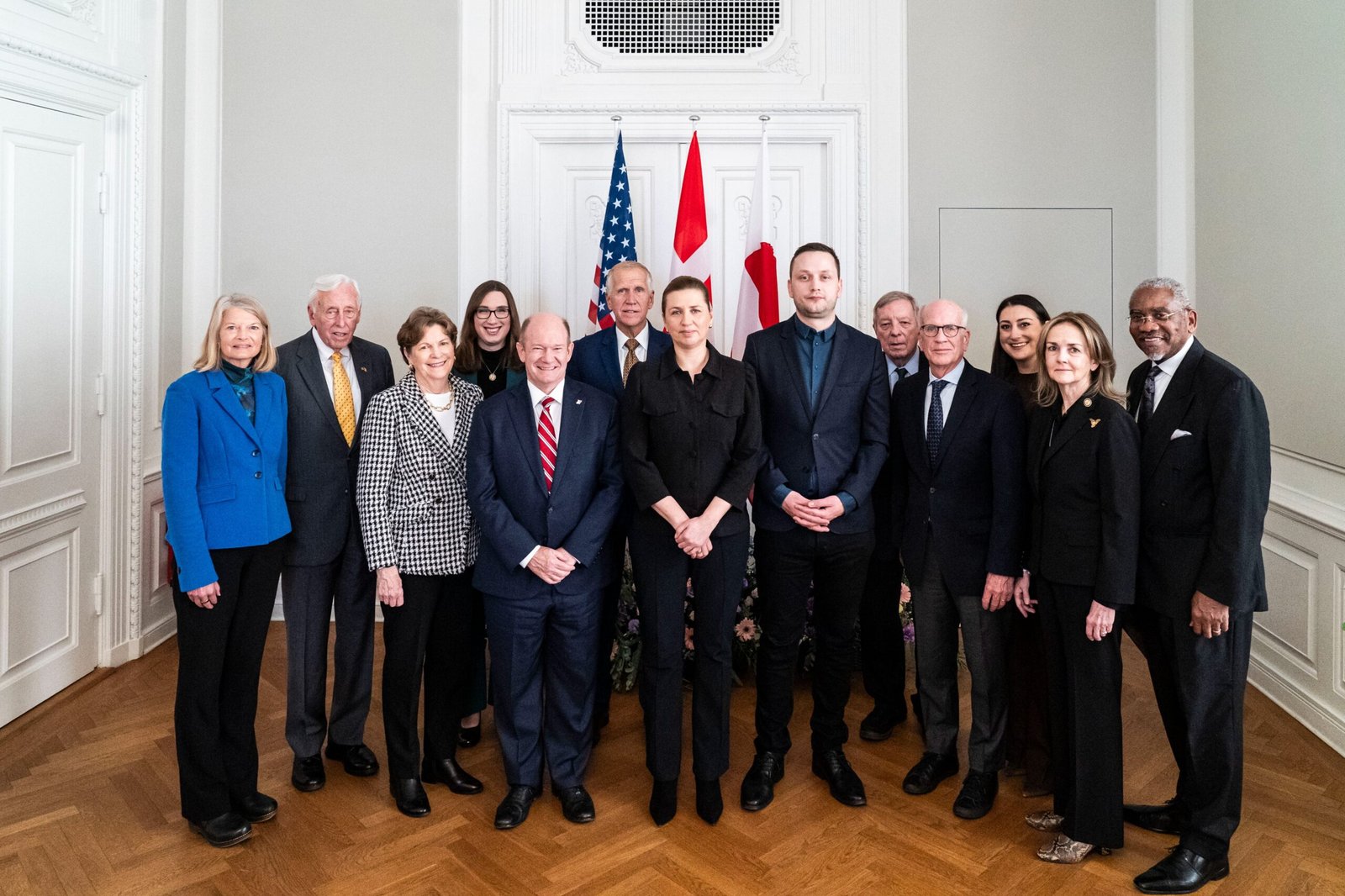 Group photo of diverse political leaders standing together in a formal setting, featuring flags of the United States and Canada in the background.