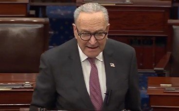 Senate Majority Leader Chuck Schumer delivering a speech on the Senate floor, wearing glasses and a suit, with an American flag pin on his lapel.
