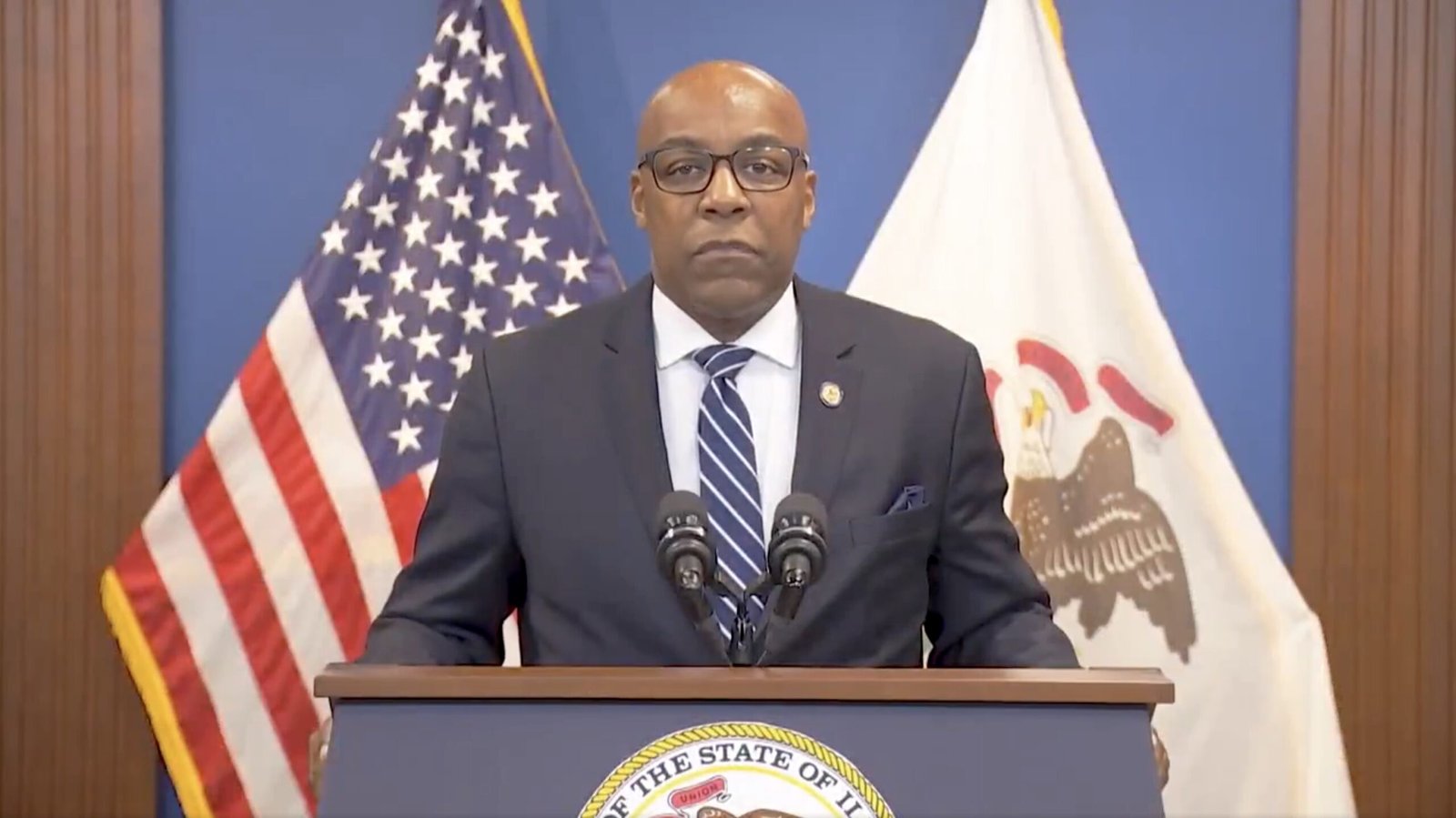 Official speaking at a podium with the American and Illinois state flags in the background, delivering a public address.
