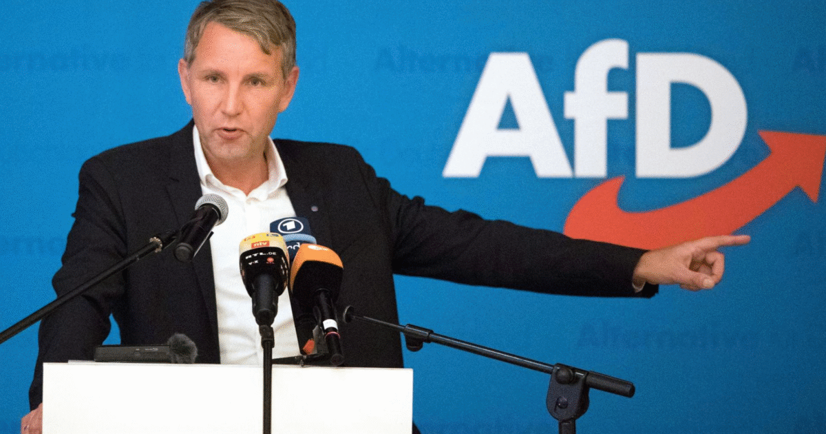 Politician speaking at a podium during an AfD event, gesturing towards the audience with a blue background featuring the AfD logo.