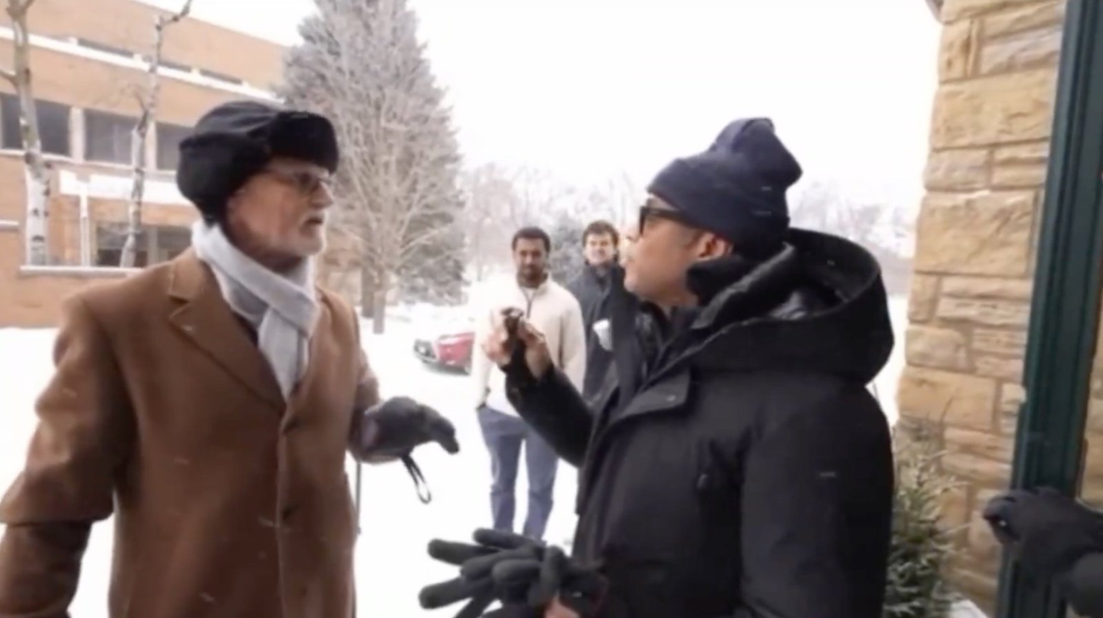 Two men engage in a conversation outside during a snowstorm, with two others observing in the background.
