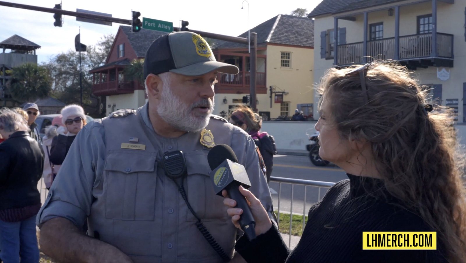 A park ranger speaks to a reporter near Fort Alley, with a crowd and historic buildings in the background.