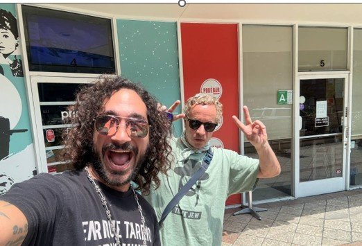 Two men posing for a selfie outside a colorful storefront, one wearing sunglasses and a black shirt, the other in a light green hoodie, both making peace signs.