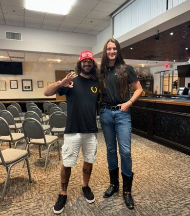 Two men pose together in a spacious indoor venue, one wearing a red cap and the other in a black shirt and cowboy boots, showcasing a casual friendship.