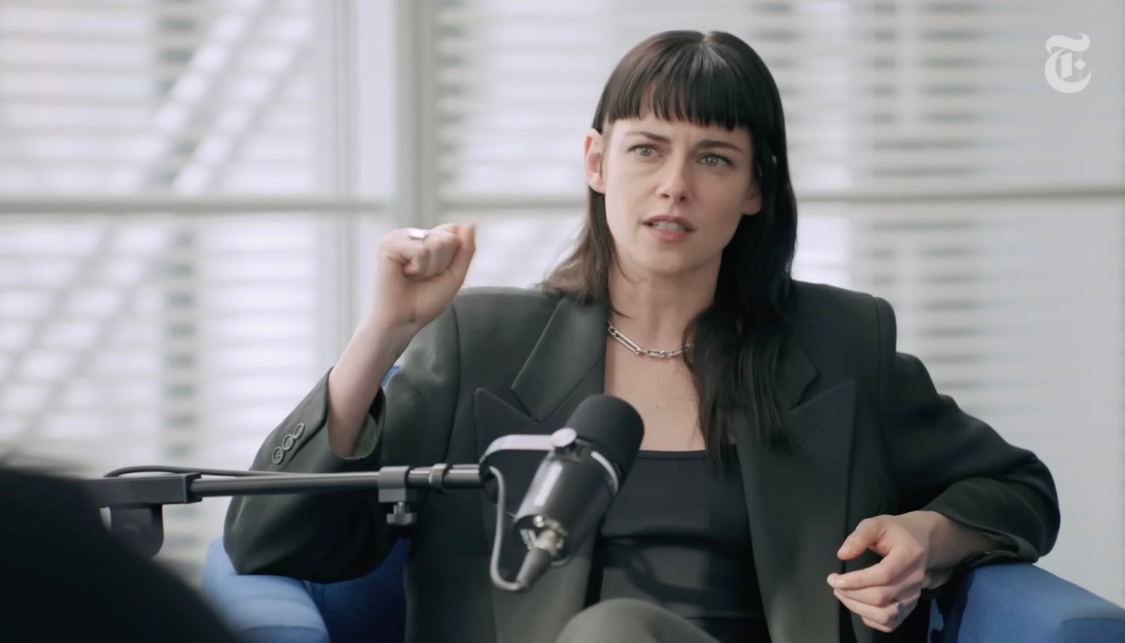 Woman in a black blazer passionately speaking during a podcast interview, with a microphone in front of her and a modern office background.