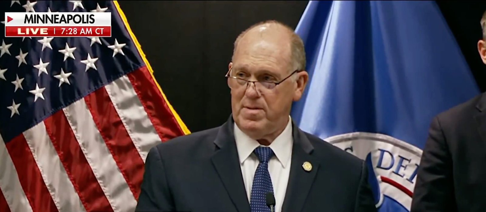 Press conference in Minneapolis featuring a speaker in a suit, with the American flag and departmental flags in the background, indicating a live broadcast.