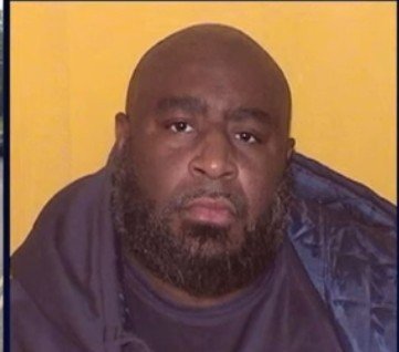 Man with a thick beard and serious expression, wearing a dark jacket against a yellow background, captured in a close-up photograph.