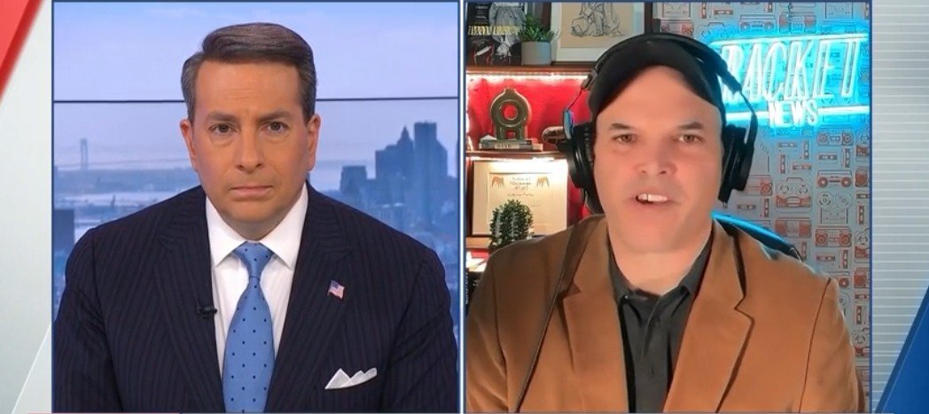 Two news anchors discussing current events, with a city skyline visible in the background and a well-decorated studio setting.