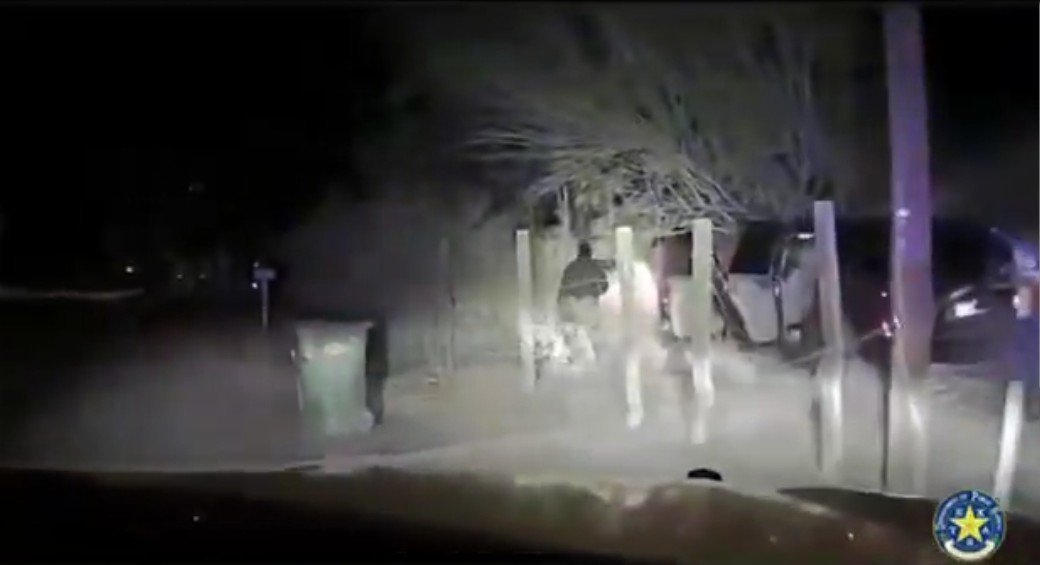 Police dashcam footage captures a nighttime scene with a figure walking near parked vehicles and a green trash can in a dimly lit area.