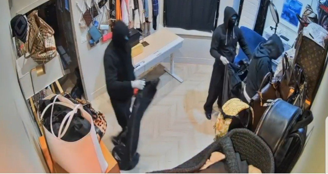 Three masked individuals in black clothing are seen stealing items from a luxury boutique, with bags and clothing scattered around the room.
